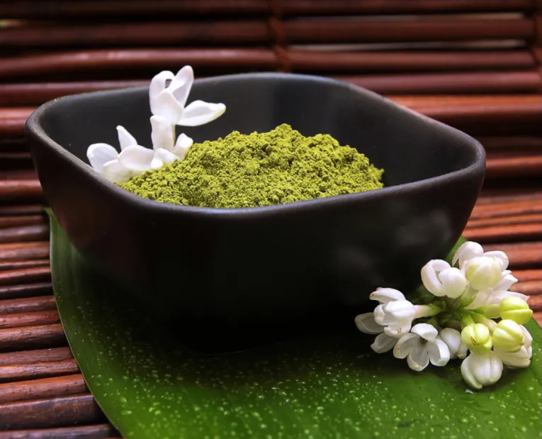 A dark bowl of fine green clay powder beside white blossoms on a leaf